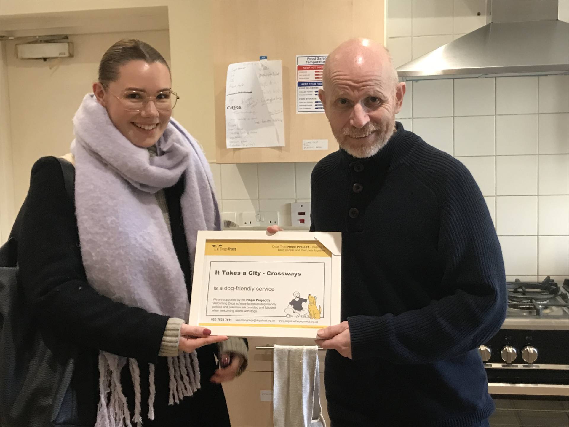 Chris from It Takes A City is standing in a kitchen receiving a certificate from a representative of Dogs Trust