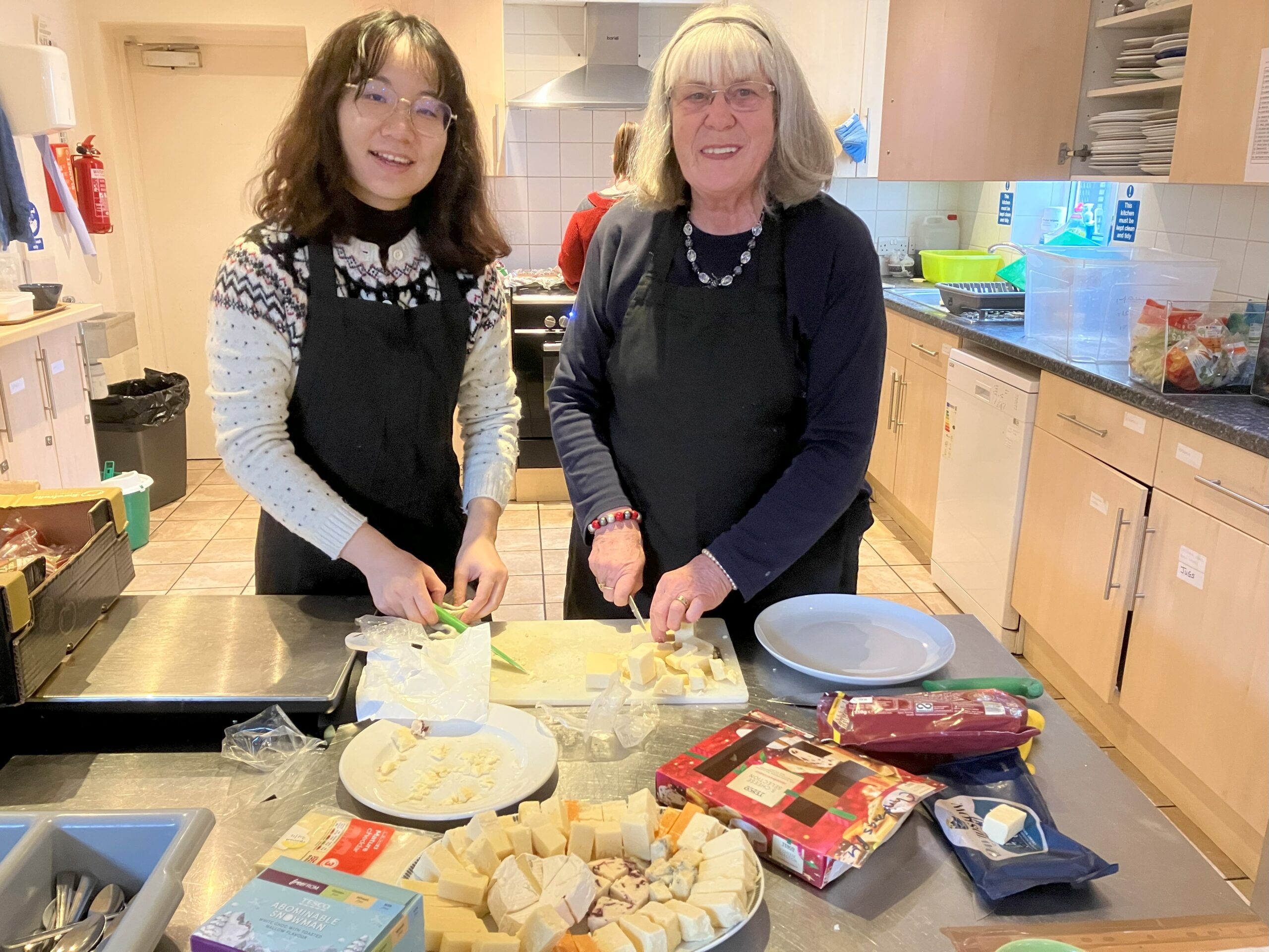 Two female volunteers look at the camera as they prepare a cheese board on a steel counter