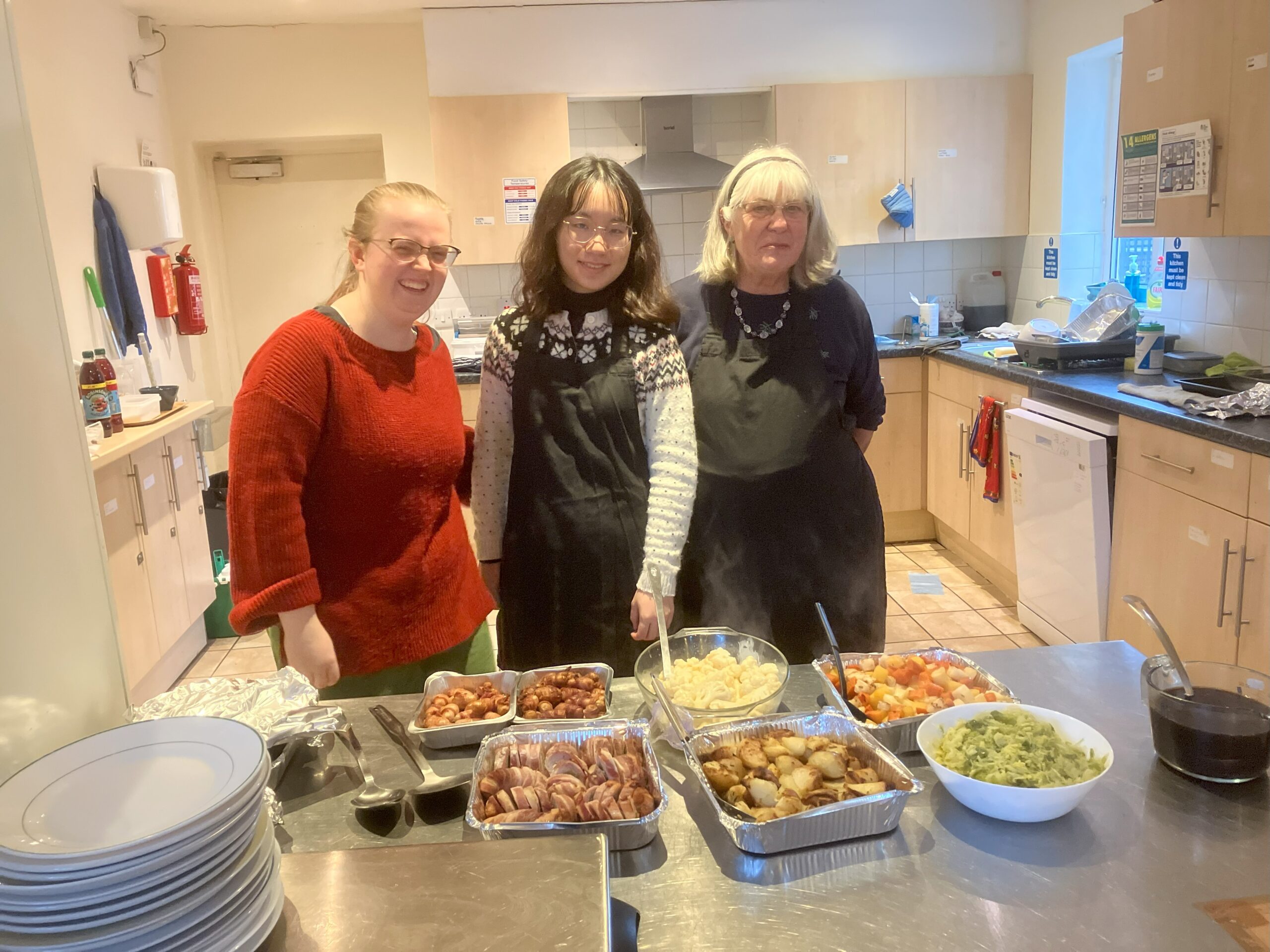 Three female volunteers stand behind a steel counter that contains serving dishes with the elements of a Christmas dinner in each.