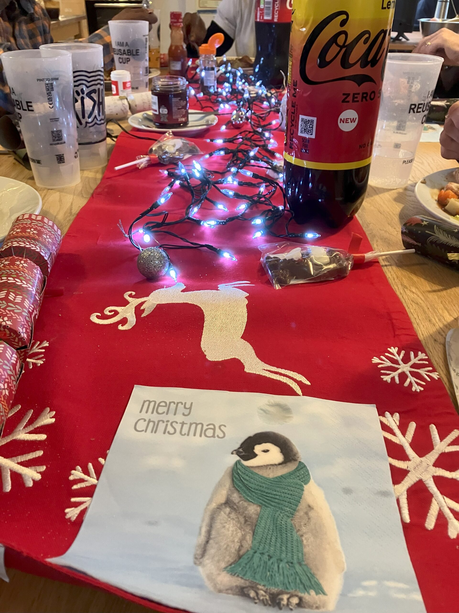 A dining table decorated with Christmas lights and tablecloth. A picture of a penguin in a scarf in the foreground