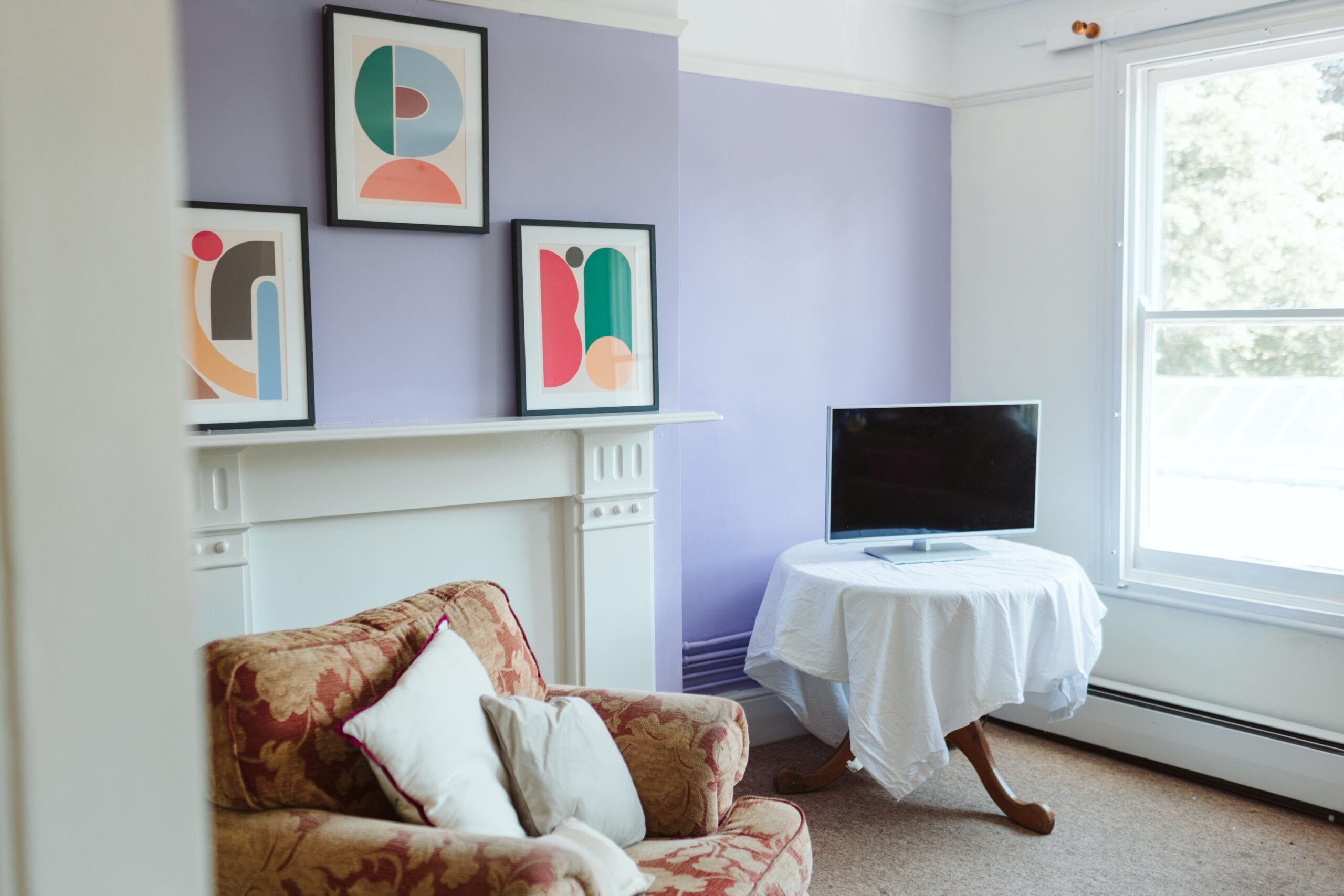 A white, moulded fire surround against a lilac wall. Three framed posters of coloured abstract designs are above the mantlepiece. In front of the fireplace is an old, comfortable armchair. To the right is a table, draped with a white tablecloth. A small TV set sits on top.