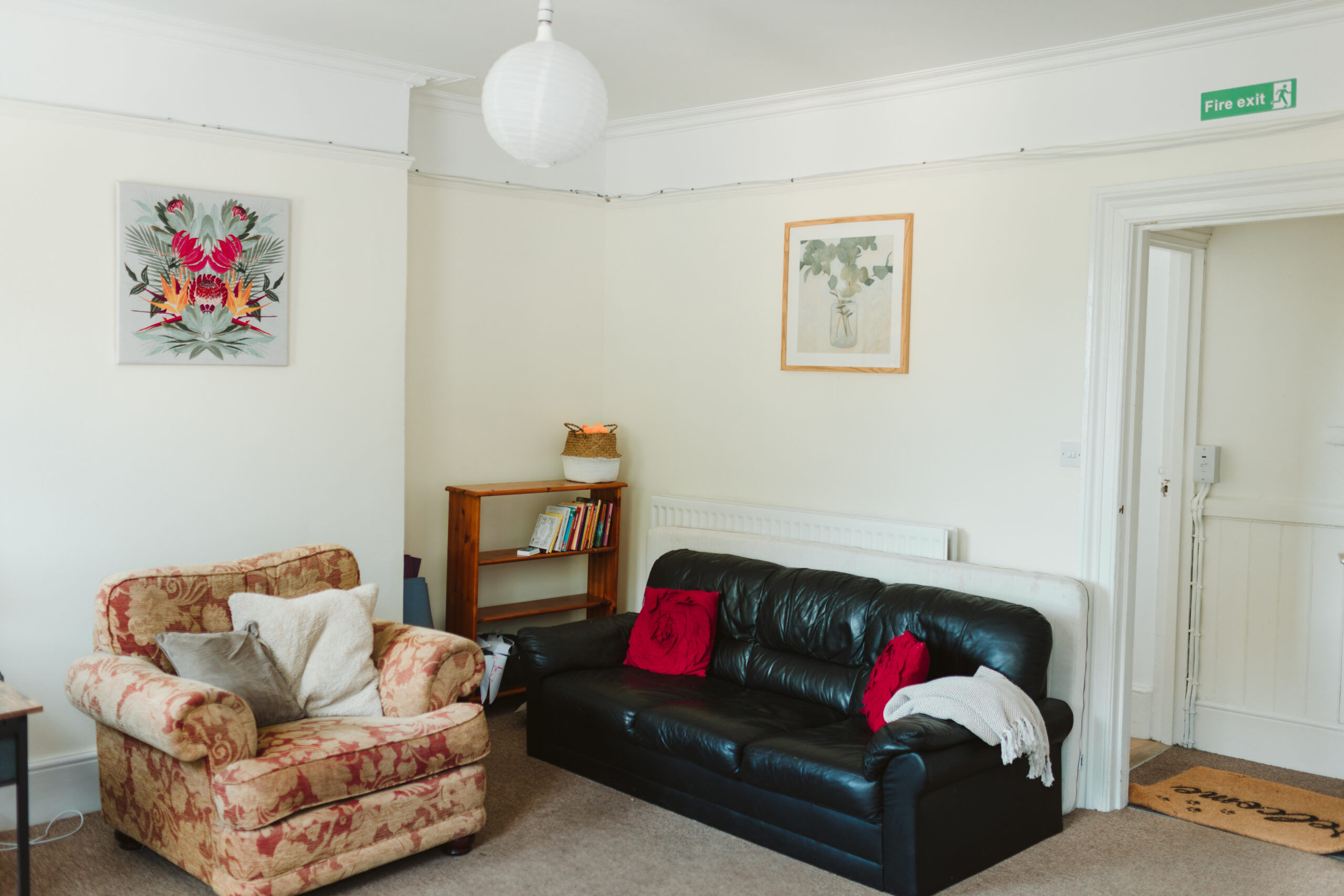 A comfortable old armchair with two large cushion sits under a floral painting on a cream wall. To the right is a three seat, black sofa with two cherry coloured cushions. A small bookcase in the background and an open door to the far right