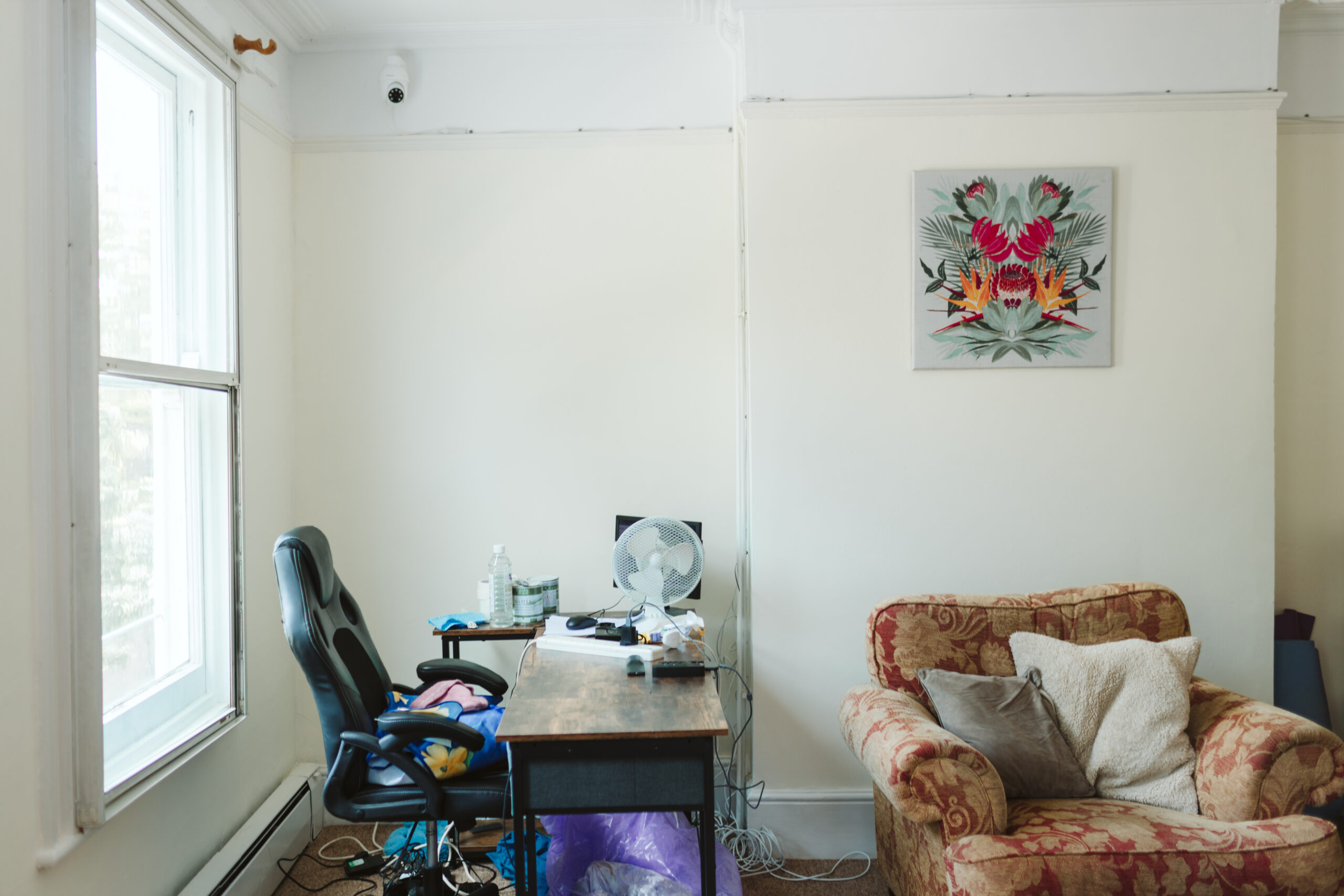 An office desk and chair sit to the left, in front of a large window. To the right is an old, comfortable armchair with two large cushions. A floral picture sits on a cream wall