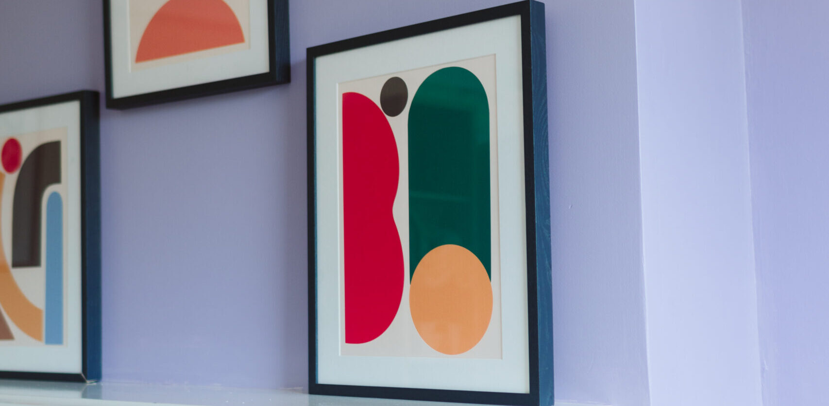 A white, moulded fire surround against a lilac wall. Three framed posters of coloured abstract designs are above the mantlepiece