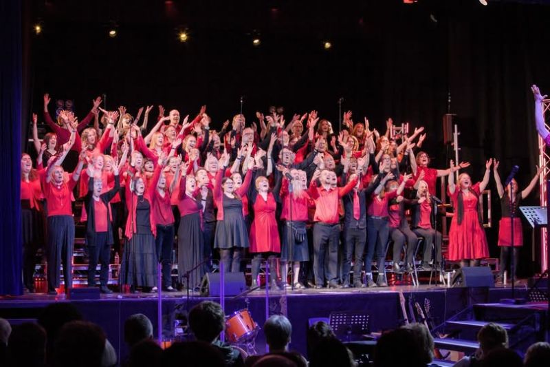 Sing Choir A large choir on a stage all holding their hands in the air and singing their hearts out. They are bathed in light