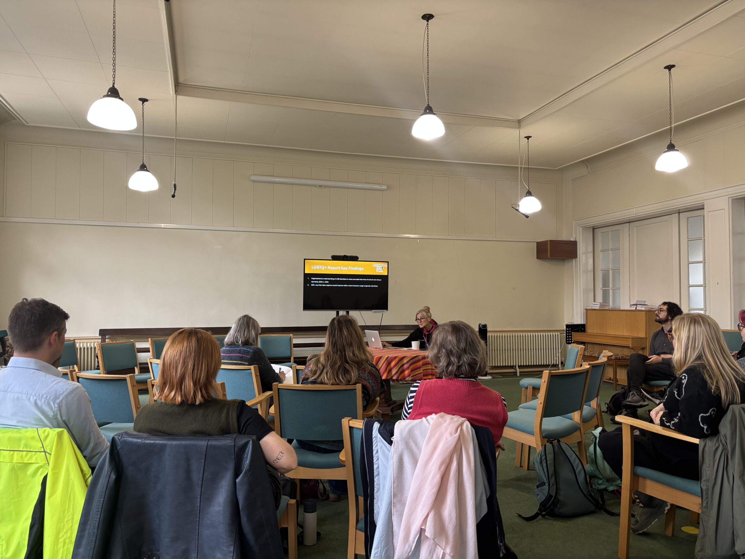 Bethan Rees from Kite Trust sits at the front of a large meeting room, delivering a training session to a group of frontline workers whose backs we can see